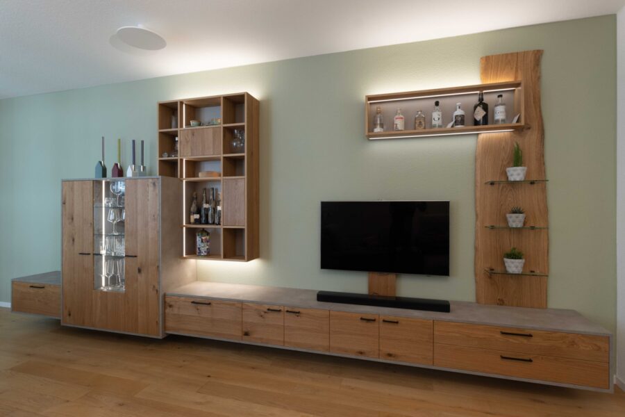 Wohnzimmer vom Schreiner: Schreinermöbel in Eiche, schwarzen Griffen, Beton-Optik und variabler LED-Beleuchtung vor salbeigrün gestrichener Wand